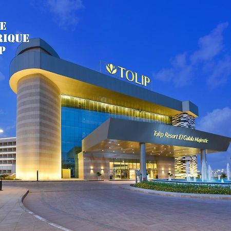 톨립 리조트 엘 갈라라 마제스틱 아인 수크나 외부 사진 The photo shows a modern hotel building named "Tolip Resort & Spa," featuring a sleek architectural design with large glass windows. The hotel is illuminated against a twilight sky, showcasing its prominent signage. In the foreground, there is a well
