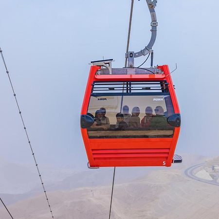 톨립 리조트 엘 갈라라 마제스틱 아인 수크나 외부 사진 The photo shows a red gondola lift suspended in the air, traveling along cables. Inside the gondola, a group of people can be seen. The background features a hazy landscape, possibly mountains or hills, indicating an elevated area. The scene conveys 