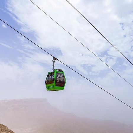 톨립 리조트 엘 갈라라 마제스틱 아인 수크나 외부 사진 The photo shows a green cable car suspended in the air, traveling along overhead cables. The backdrop features a hazy landscape, likely mountainous or elevated terrain, with a cloudy sky. The scene suggests a scenic transportation method, possibly us