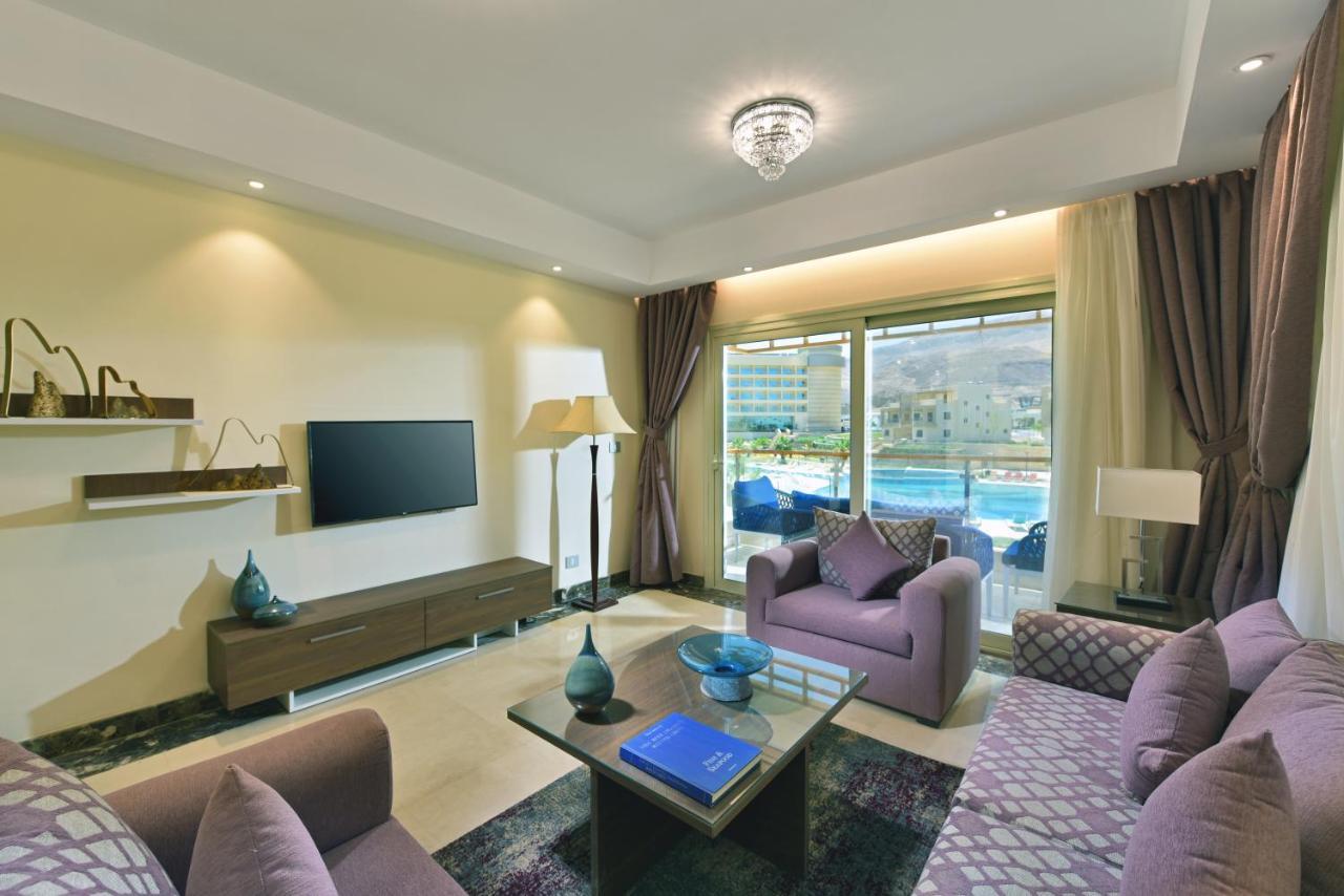 톨립 리조트 엘 갈라라 마제스틱 아인 수크나 외부 사진 The photo shows a modern living room that features a stylish and cozy design. There are two purple sofas with decorative cushions arranged around a coffee table. The coffee table has a blue book and a decorative item on it. A flat-screen TV is mounte