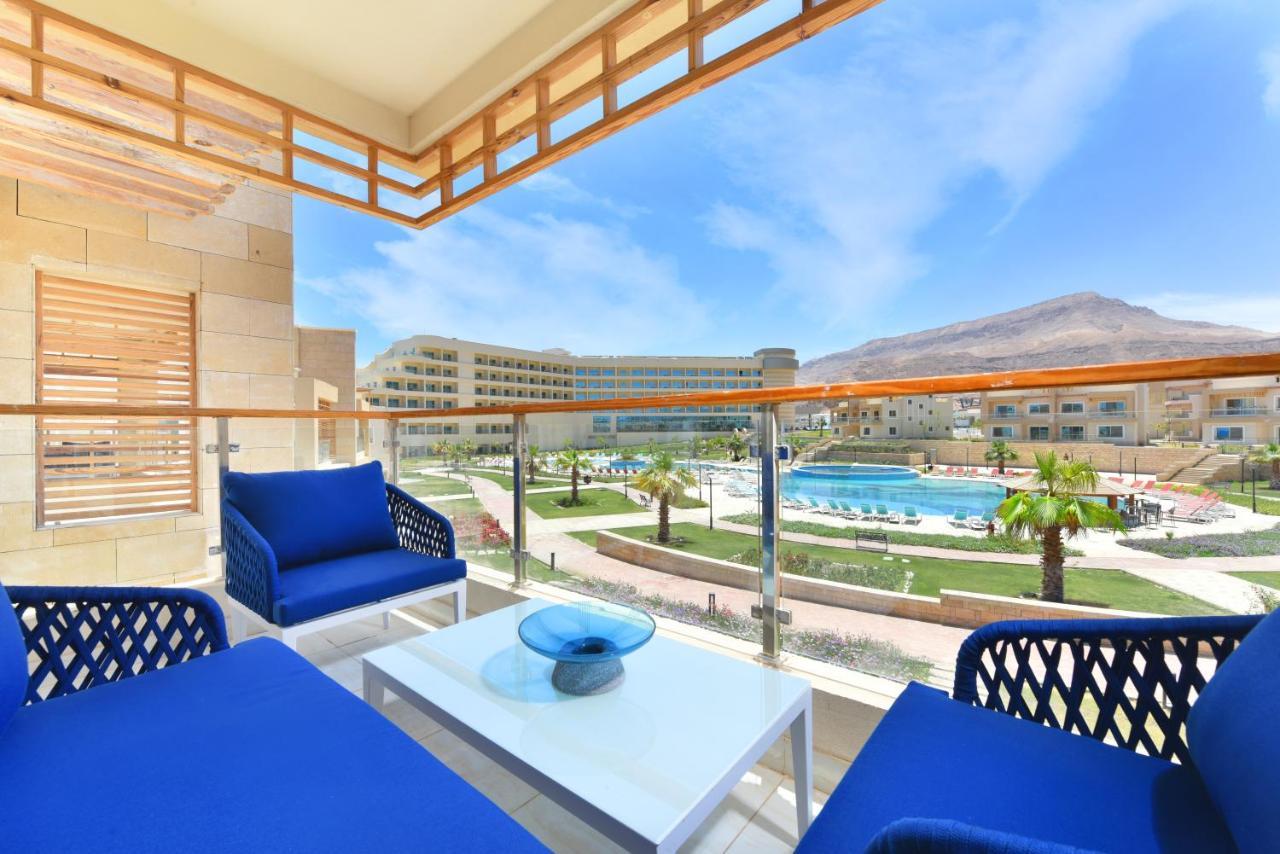 톨립 리조트 엘 갈라라 마제스틱 아인 수크나 외부 사진 The photo showcases a balcony view featuring two blue chairs and a table with a decorative bowl. The balcony overlooks a landscape that includes a swimming pool and manicured lawns. In the background, there are multiple buildings and palm trees, all 