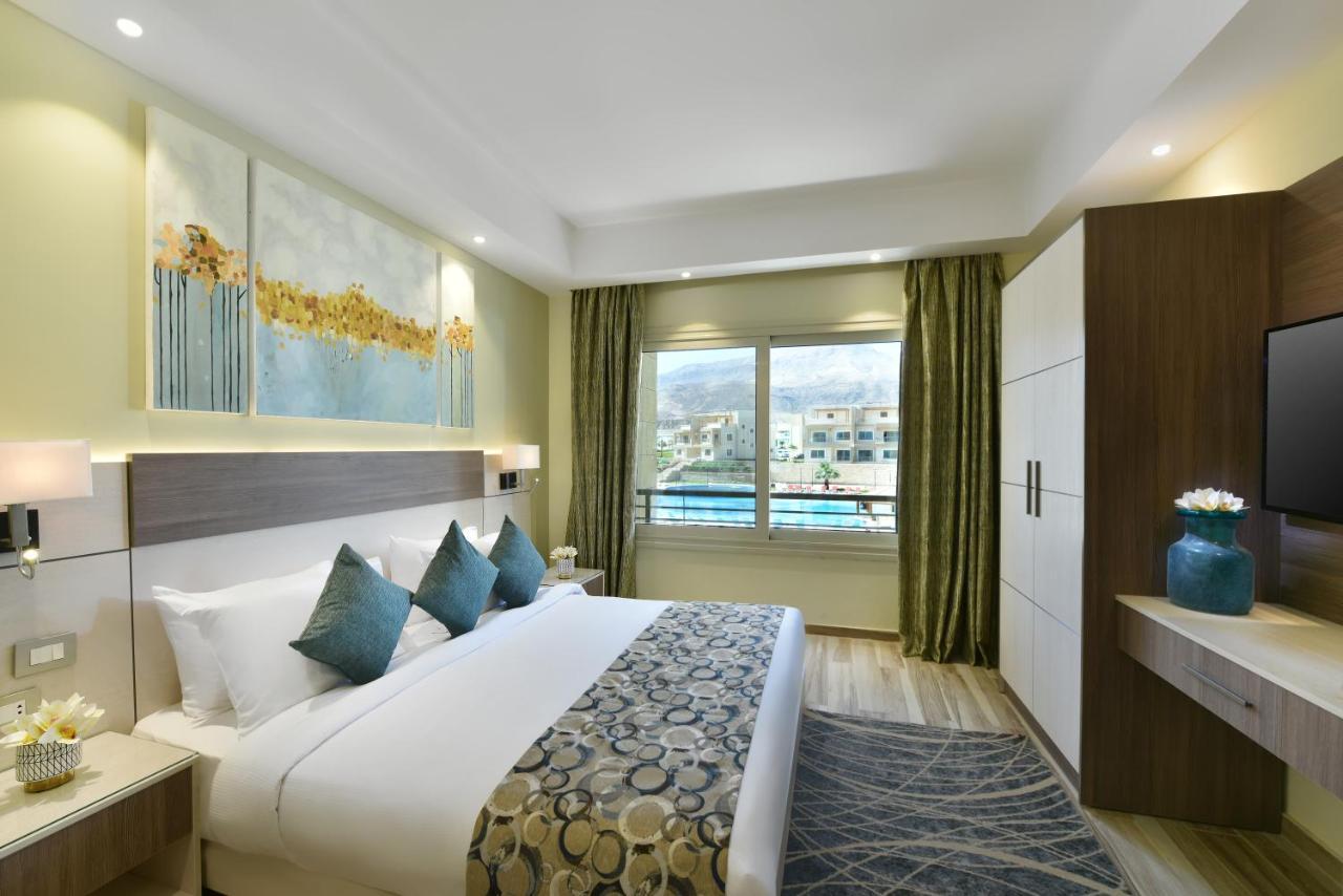 톨립 리조트 엘 갈라라 마제스틱 아인 수크나 외부 사진 The photo shows a modern hotel room. It features a large bed with white bedding and several decorative pillows in shades of teal. Above the bed, there are two pieces of wall art with a landscape theme. The room has a large window with green curtains,