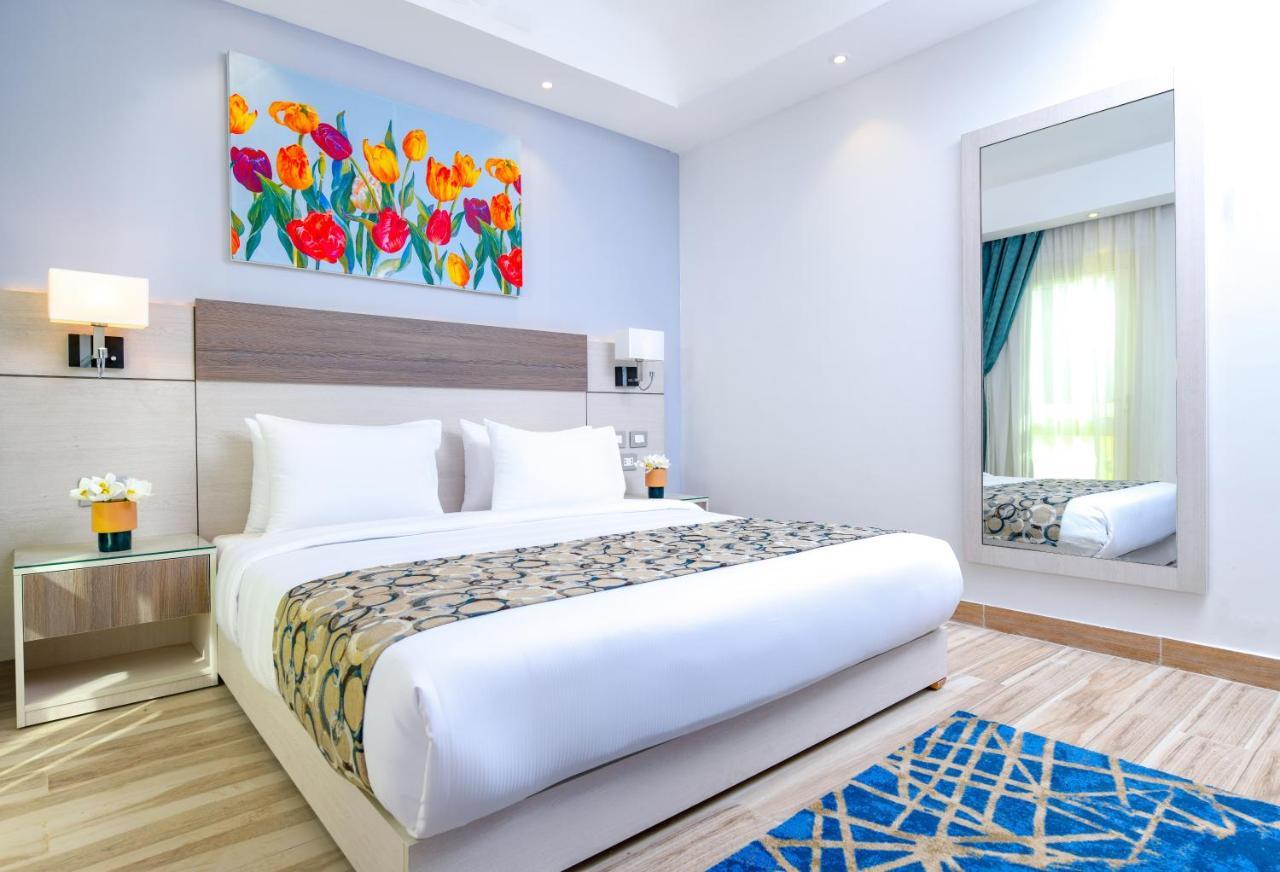 톨립 리조트 엘 갈라라 마제스틱 아인 수크나 외부 사진 The photo shows a modern hotel bedroom. It features a large bed with white linens and a decorative throw blanket. The room has light wooden flooring and is bright, with ample natural light coming from a window. There is a colorful painting of flowers