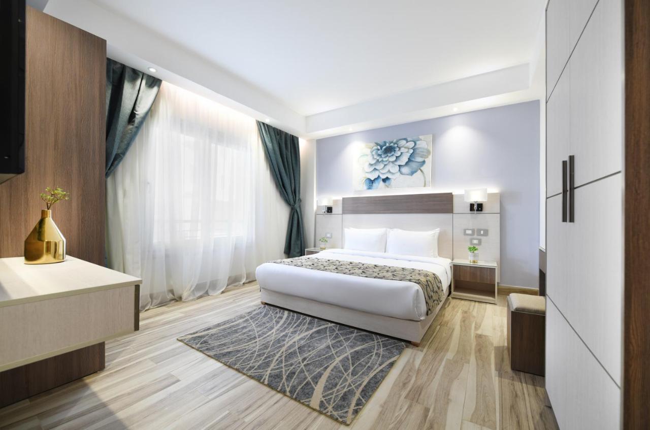 톨립 리조트 엘 갈라라 마제스틱 아인 수크나 외부 사진 The photo shows a modern bedroom with a clean and stylish design. It features a large bed with white bedding positioned against a wall. Above the bed, there is a decorative artwork, possibly a floral design. The room has large windows with sheer curt