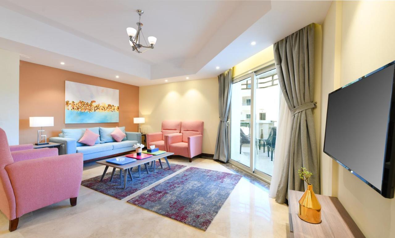 톨립 리조트 엘 갈라라 마제스틱 아인 수크나 객실 사진 The photo shows a modern living room that features a cozy and stylish design. There is a light blue sofa adorned with pastel pink cushions, accompanied by two pink armchairs. A coffee table sits in front, placed on a colorful area rug that adds warmt