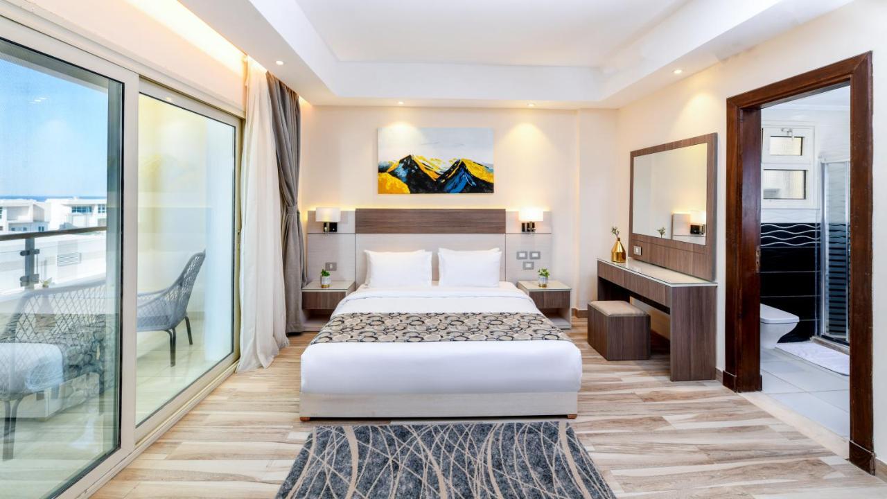톨립 리조트 엘 갈라라 마제스틱 아인 수크나 외부 사진 The photo shows a modern hotel room featuring a large bed with white linens and a decorative throw. The room has a neutral color palette, accented with warm lighting. There is a piece of artwork above the bed depicting mountains. To the left, there a