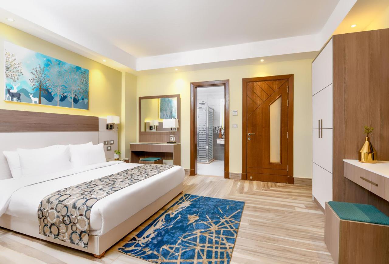 톨립 리조트 엘 갈라라 마제스틱 아인 수크나 외부 사진 The photo shows a well-decorated hotel room. There is a large bed with white linens and a decorative throw blanket. The room features a wooden wardrobe and a bedside table with lamps. A colorful rug is placed on the floor, adding warmth to the space.