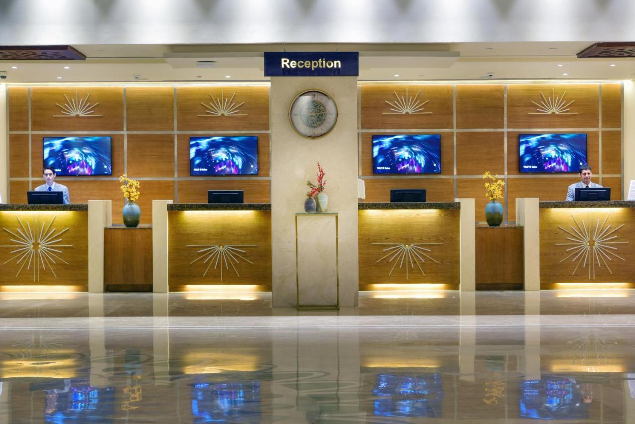 톨립 리조트 엘 갈라라 마제스틱 아인 수크나 외부 사진 The image shows a hotel reception area. There are several reception desks lined up against a wall with decorative paneling. Each desk has a staff member, and above the desks, there are screens displaying information. A clock is visible on the wall, a