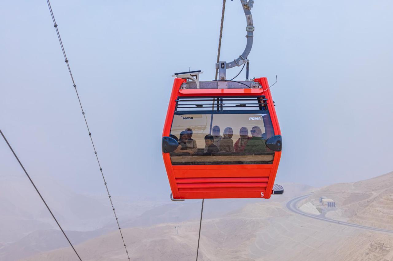 톨립 리조트 엘 갈라라 마제스틱 아인 수크나 외부 사진 The photo shows a red gondola lift suspended in the air, traveling along cables. Inside the gondola, a group of people can be seen. The background features a hazy landscape, possibly mountains or hills, indicating an elevated area. The scene conveys 