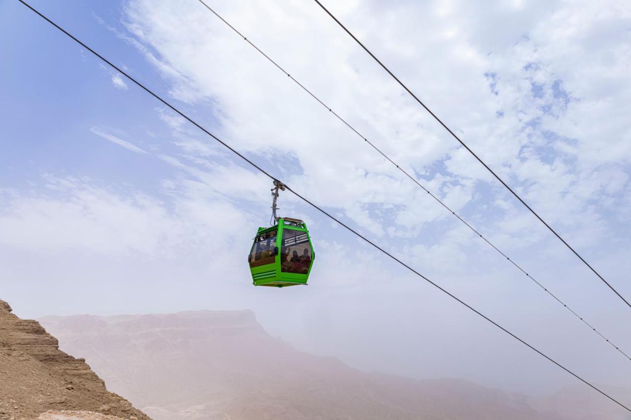 톨립 리조트 엘 갈라라 마제스틱 아인 수크나 외부 사진 The photo shows a green cable car suspended in the air, traveling along overhead cables. The backdrop features a hazy landscape, likely mountainous or elevated terrain, with a cloudy sky. The scene suggests a scenic transportation method, possibly us