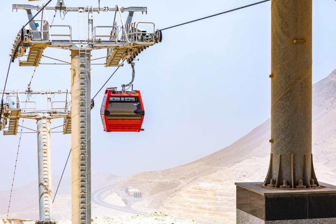 톨립 리조트 엘 갈라라 마제스틱 아인 수크나 외부 사진 The photo shows a cable car system with a single red gondola suspended mid-air. It appears to be part of a larger cable car infrastructure, with metal pylons supporting the cables above. The background features a hazy landscape, likely characterized 