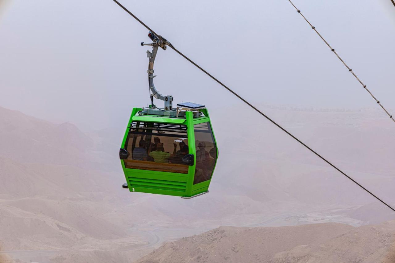 톨립 리조트 엘 갈라라 마제스틱 아인 수크나 외부 사진 The photo shows a green cable car suspended in the air, moving along a wire. The cable car appears to be traversing a mountainous landscape, which looks rocky and arid. The surrounding environment is somewhat foggy or hazy, suggesting limited visibil