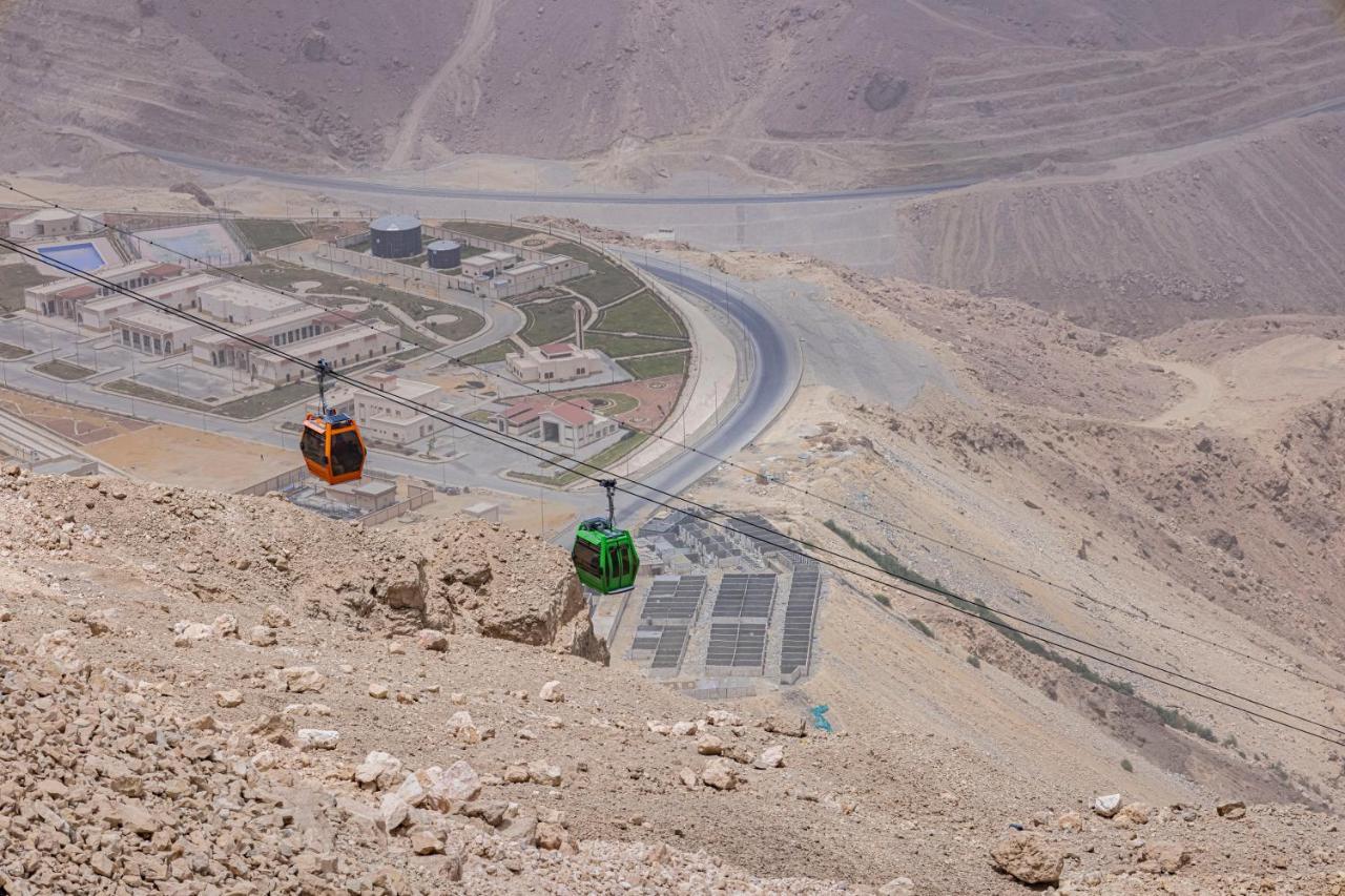 톨립 리조트 엘 갈라라 마제스틱 아인 수크나 외부 사진 The photo shows a scenic view from a high elevation, featuring a cable car system. Two cable cars are visible, one orange and one green, traveling along cables down the mountainside. Below, there appears to be a developed area with roads and light st