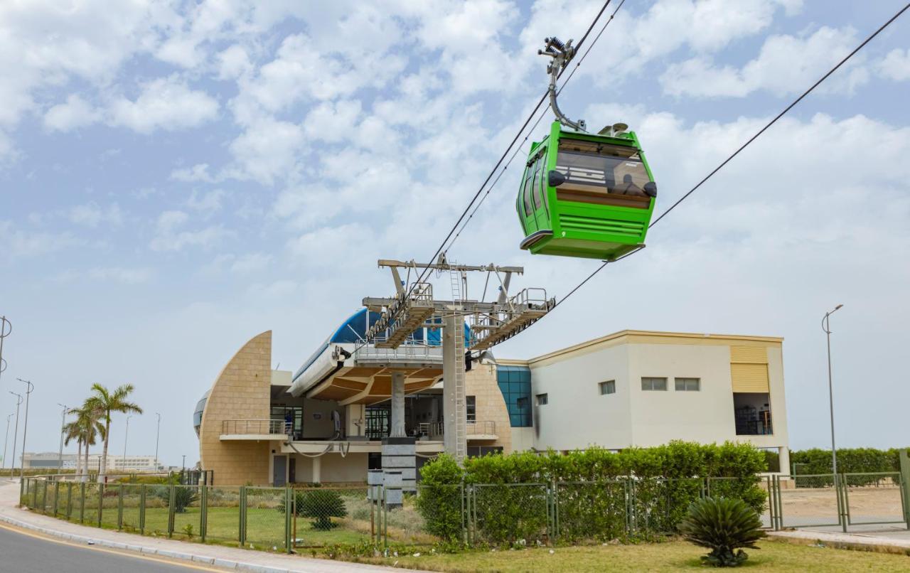 톨립 리조트 엘 갈라라 마제스틱 아인 수크나 외부 사진 The photo shows a cable car or gondola system in operation, featuring a green-colored gondola. It is suspended from cables and moving towards a station, which appears to be a modern building with a unique architectural design. The station has large w