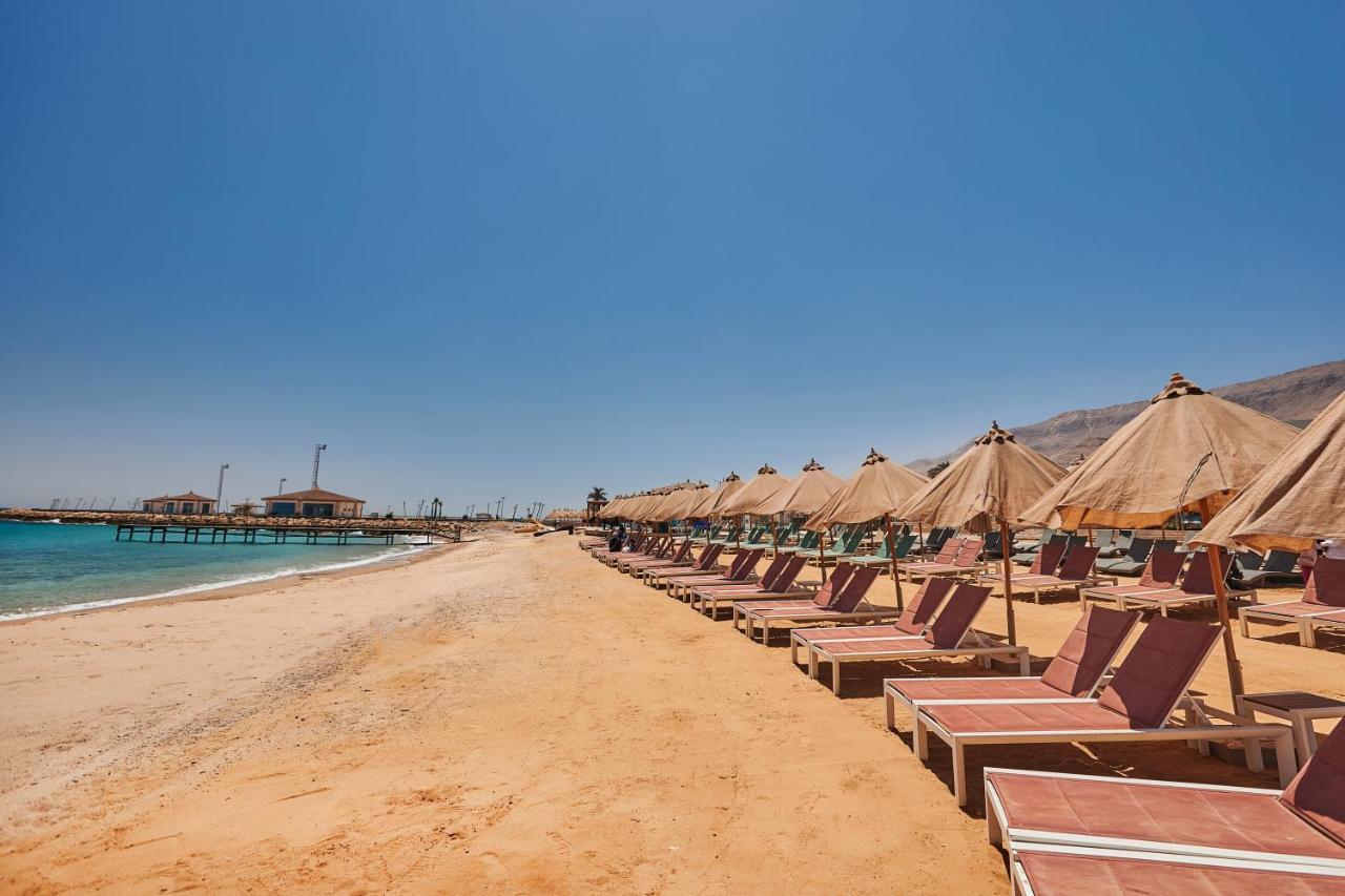 톨립 리조트 엘 갈라라 마제스틱 아인 수크나 외부 사진 The photo shows a scenic beach with a stretch of golden sand under a clear blue sky. There are several beach loungers arranged in rows, each accompanied by thatched umbrellas providing shade. In the background, there is a wooden pier extending into t