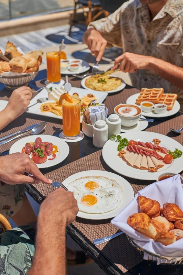 톨립 리조트 엘 갈라라 마제스틱 아인 수크나 외부 사진 The photo features a spread of breakfast dishes on a table. There are two sunny-side-up eggs on one plate, surrounded by various food items, including slices of ham, sausages, and fresh tomatoes. Another plate holds a serving of waffles. A bowl with 