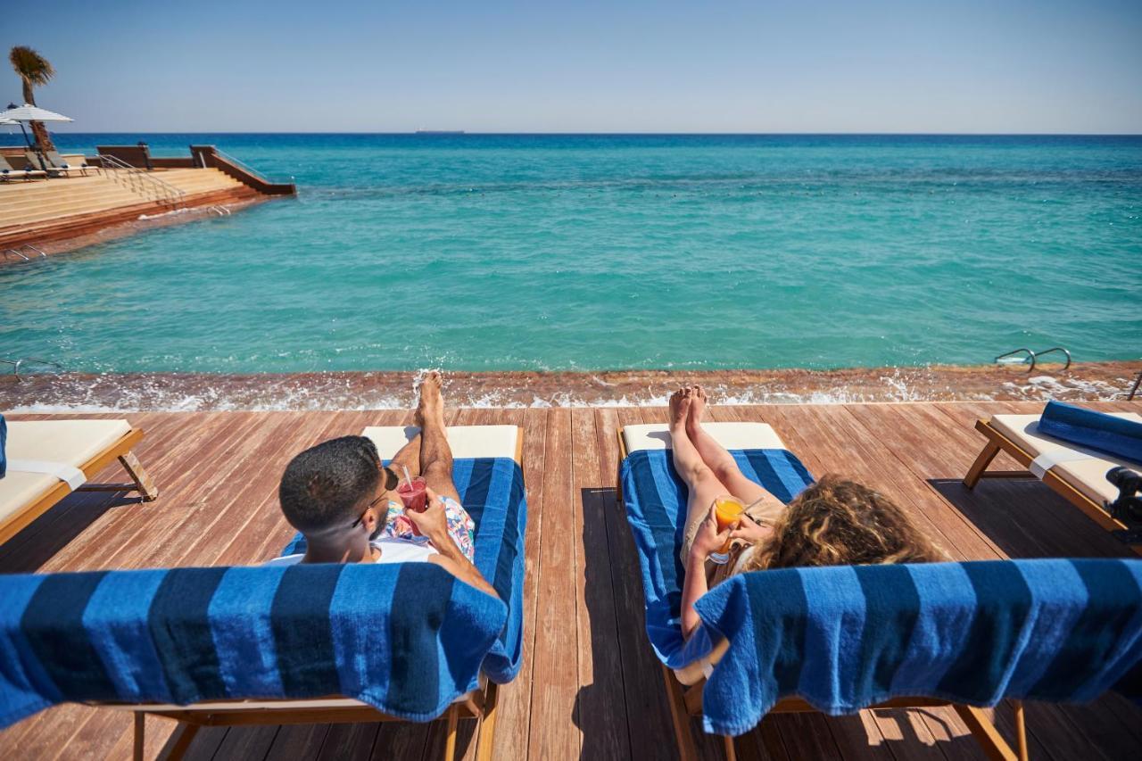 톨립 리조트 엘 갈라라 마제스틱 아인 수크나 외부 사진 The photo depicts two people relaxing on lounge chairs by the waterfront. They are positioned on a wooden deck with a view of the clear, turquoise water. Both individuals are reclining, enjoying the sun, and each has a drink in hand. The scene convey