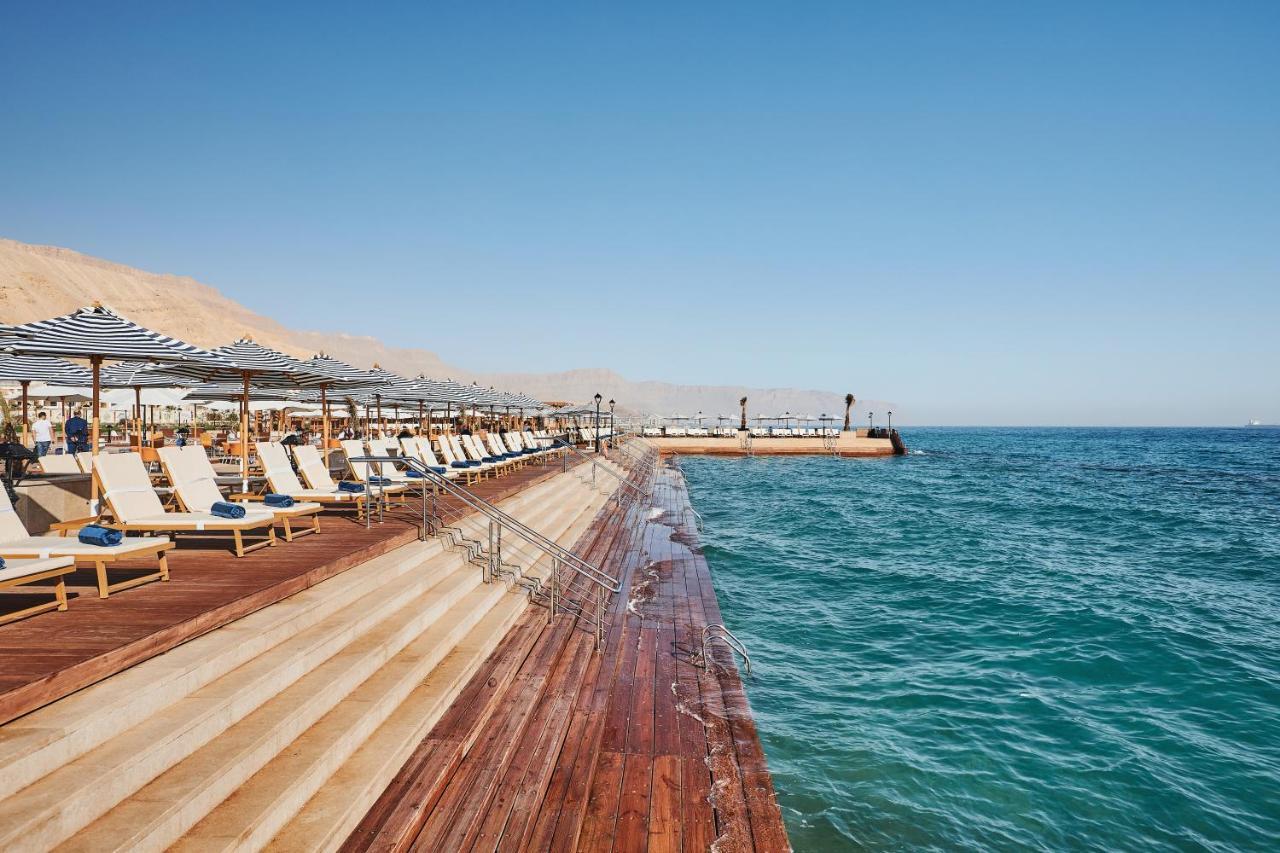 톨립 리조트 엘 갈라라 마제스틱 아인 수크나 외부 사진 The photo shows a seaside area featuring a wooden deck that extends into the water. On the left side, there are several lounge chairs arranged neatly under shade structures, suggesting a relaxing atmosphere. The ocean is visible in the foreground, wi