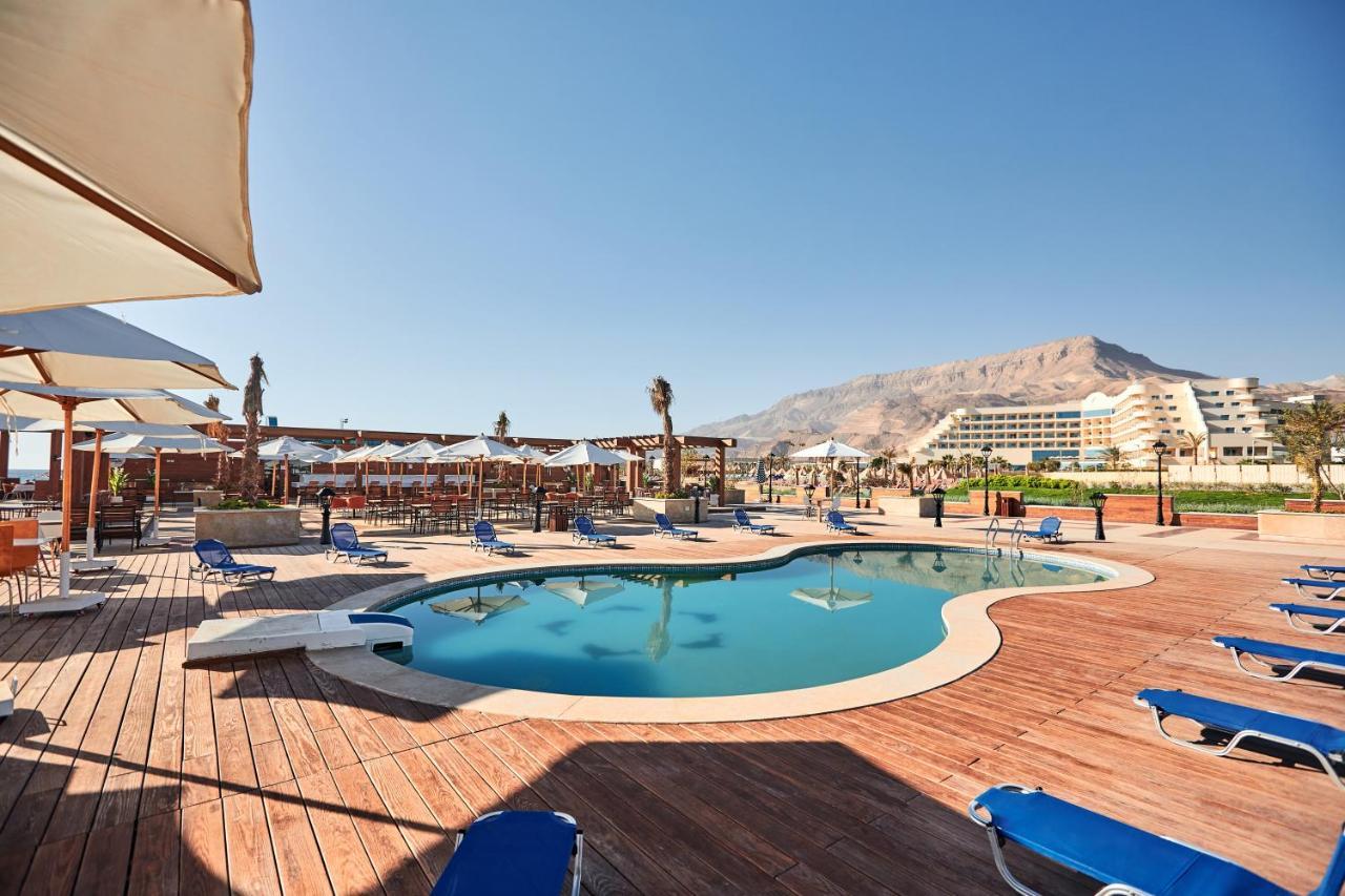 톨립 리조트 엘 갈라라 마제스틱 아인 수크나 외부 사진 The photo shows a swimming pool area surrounded by sun loungers and umbrellas. The pool has a unique, curvy shape and is situated on a wooden deck. In the background, there are several buildings, likely part of a resort or hotel, and a mountainous la