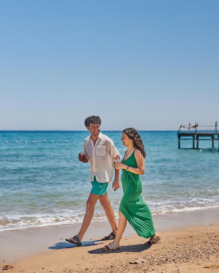 톨립 리조트 엘 갈라라 마제스틱 아인 수크나 외부 사진 The photo shows a couple walking along a beach. The man is wearing a white short-sleeved shirt and green shorts, while the woman is dressed in a flowing green dress. They both appear to be enjoying the pleasant weather, with the ocean in the backgrou