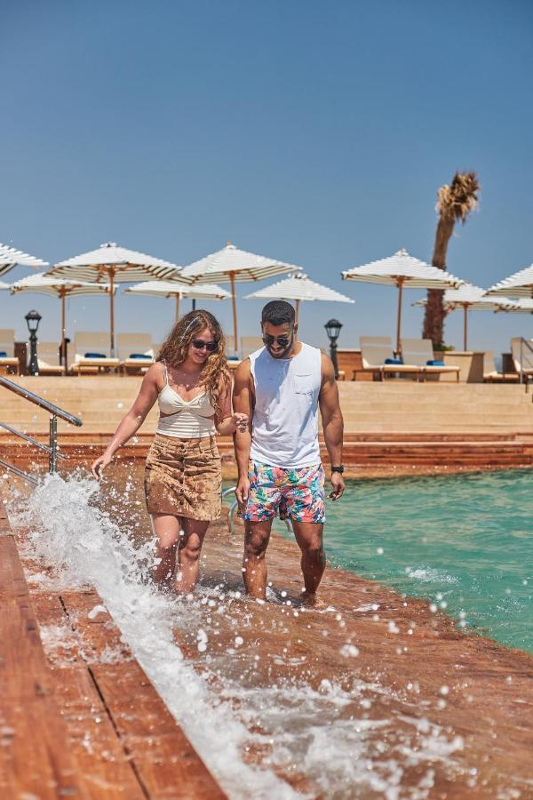 톨립 리조트 엘 갈라라 마제스틱 아인 수크나 외부 사진 The photo depicts a couple walking along a poolside area. They are enjoying a sunny day near a swimming pool, with splashes of water around them. The woman is wearing a light-colored top and a patterned skirt, while the man is dressed in a sleeveless