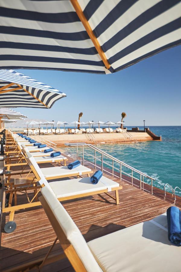 톨립 리조트 엘 갈라라 마제스틱 아인 수크나 외부 사진 The photo depicts a serene outdoor area by the waterfront. There are several lounge chairs arranged in a row, each equipped with blue towels. Large, striped umbrellas provide shade over some of the chairs. In the background, there is a wooden deck th