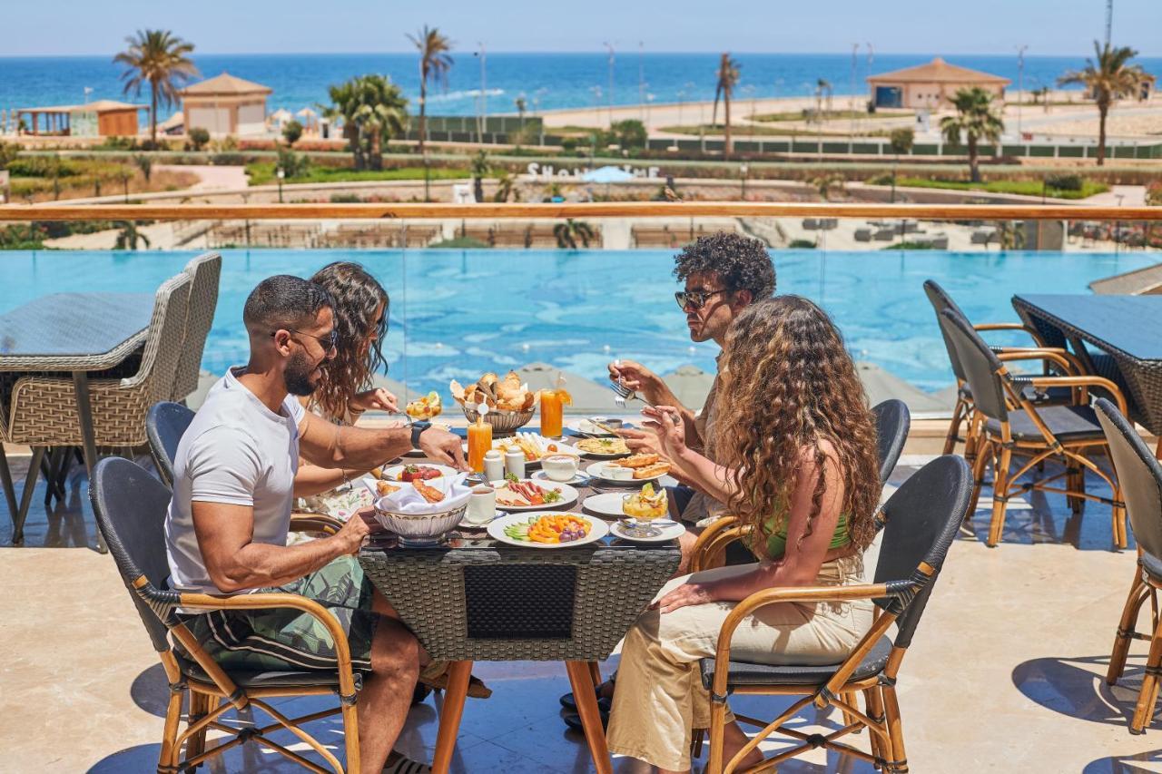 톨립 리조트 엘 갈라라 마제스틱 아인 수크나 외부 사진 The photo shows a group of five people enjoying a meal together outdoors, likely at a restaurant or cafe. They are seated at a table with a beautiful view of a swimming pool and the sea in the background. The table is filled with various dishes, and 