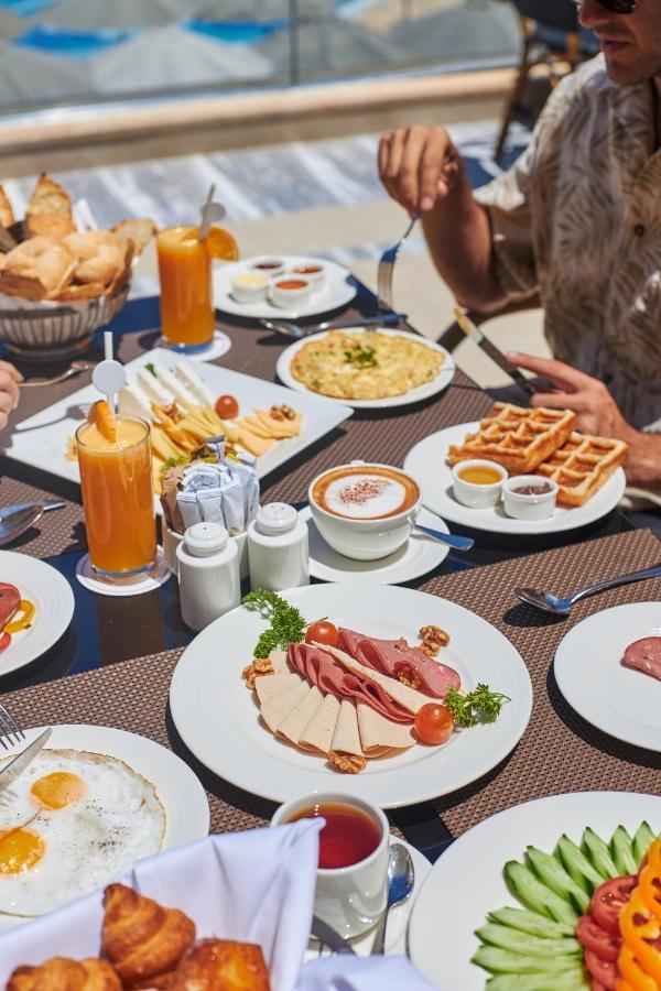 톨립 리조트 엘 갈라라 마제스틱 아인 수크나 외부 사진 The photo shows a breakfast spread laid out on a table. There are several plates with a variety of food items, including:

1. **Sliced Cold Cuts**: A plate featuring various cold meats, accompanied by garnishes like cherry tomatoes and lettuce.
2. **