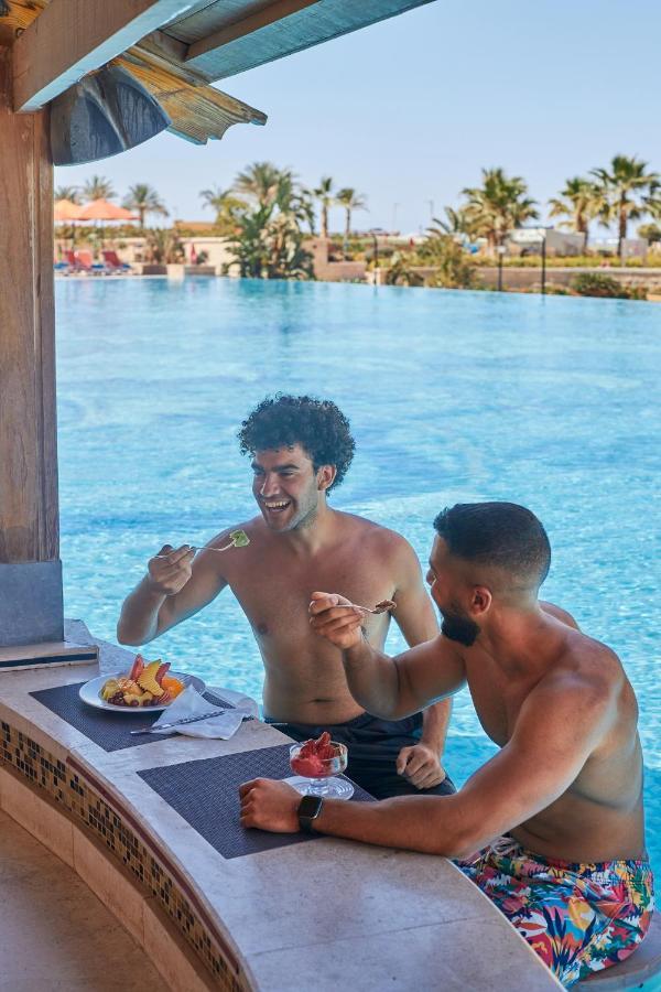 톨립 리조트 엘 갈라라 마제스틱 아인 수크나 외부 사진 The photo features two young men enjoying a meal together. They are sitting at a poolside table with a clear blue swimming pool in the background. The men are shirtless, suggesting a casual, relaxed atmosphere, likely in a warm environment. One man i