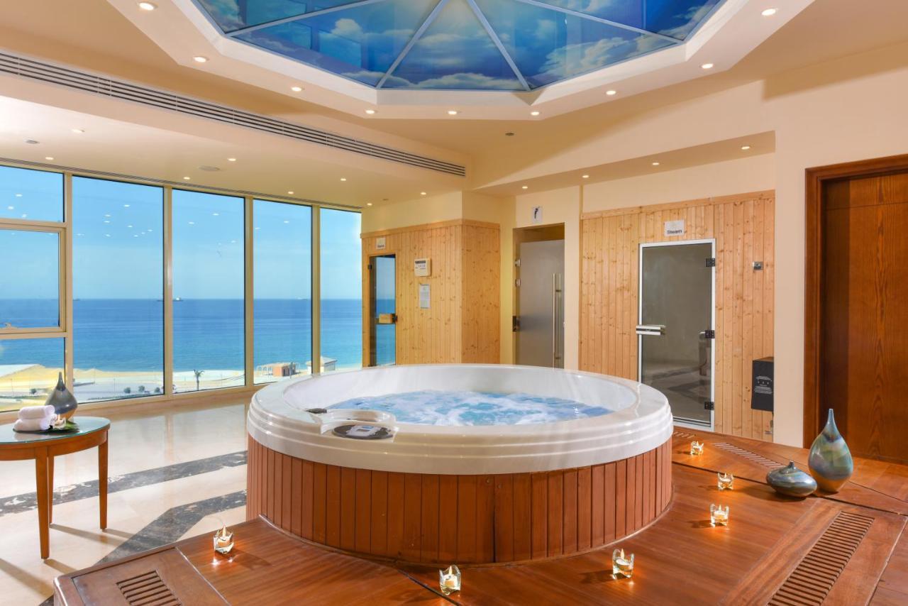 톨립 리조트 엘 갈라라 마제스틱 아인 수크나 외부 사진 The photo shows a luxurious spa room featuring a large, round hot tub positioned in the center. The walls are mostly glass, offering a stunning view of the ocean. The ceiling has a painted sky, enhancing the serene atmosphere. There are wooden elemen