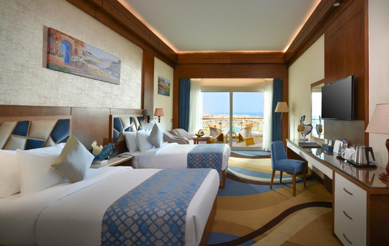 톨립 리조트 엘 갈라라 마제스틱 아인 수크나 외부 사진 The photo shows a well-decorated hotel room featuring two beds with decorative pillows. The room has wooden accents and modern furnishings, including a desk, a television, and lamps. Large windows flood the space with natural light, offering a view o