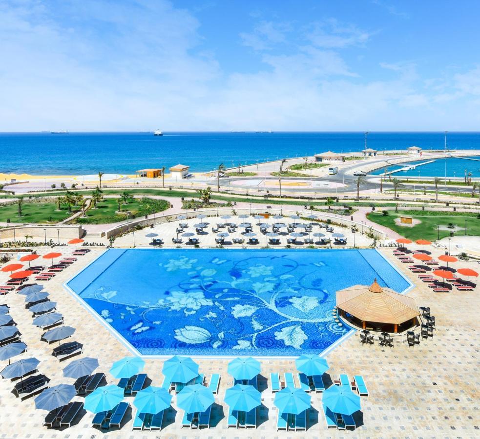 톨립 리조트 엘 갈라라 마제스틱 아인 수크나 외부 사진 The photo features a large swimming pool with a decorative design on its surface, surrounded by sun loungers and umbrellas. In the background, there is a view of the sea, with a clear blue sky and a few scattered clouds. The area is well-maintained, 