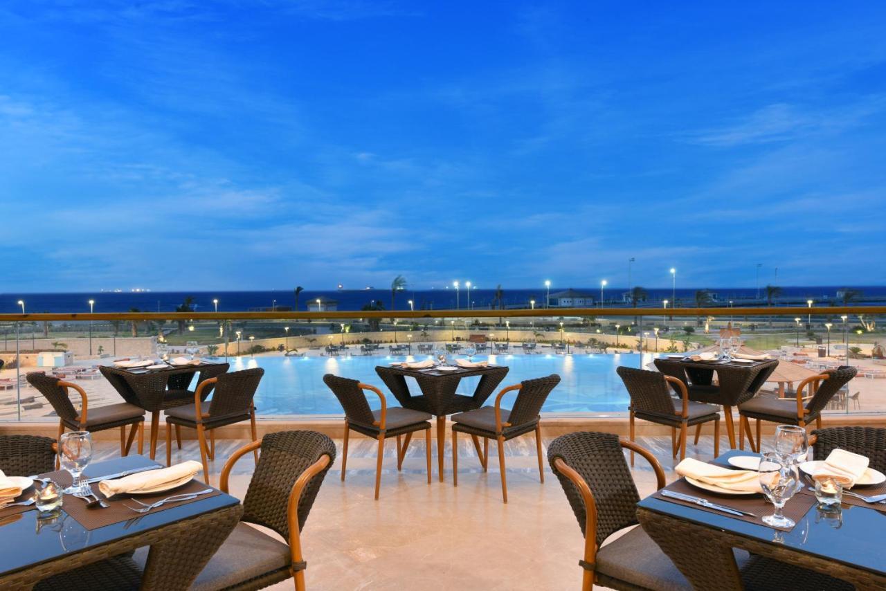 톨립 리조트 엘 갈라라 마제스틱 아인 수크나 외부 사진 The photo shows an outdoor dining area with several neatly arranged tables and chairs. The tables are elegantly set with cutlery and plates, suggesting a fine dining experience. In the background, there is a large swimming pool that reflects the blue