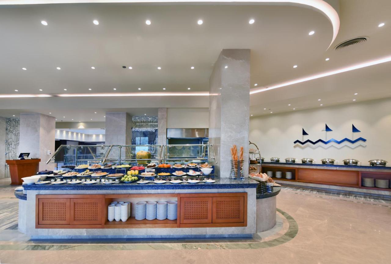 톨립 리조트 엘 갈라라 마제스틱 아인 수크나 외부 사진 The image shows a buffet-style dining area in a restaurant or hotel. There are several food stations with a variety of dishes displayed, including fruits, salads, and other food items. The layout features a large, central island with a wooden base an