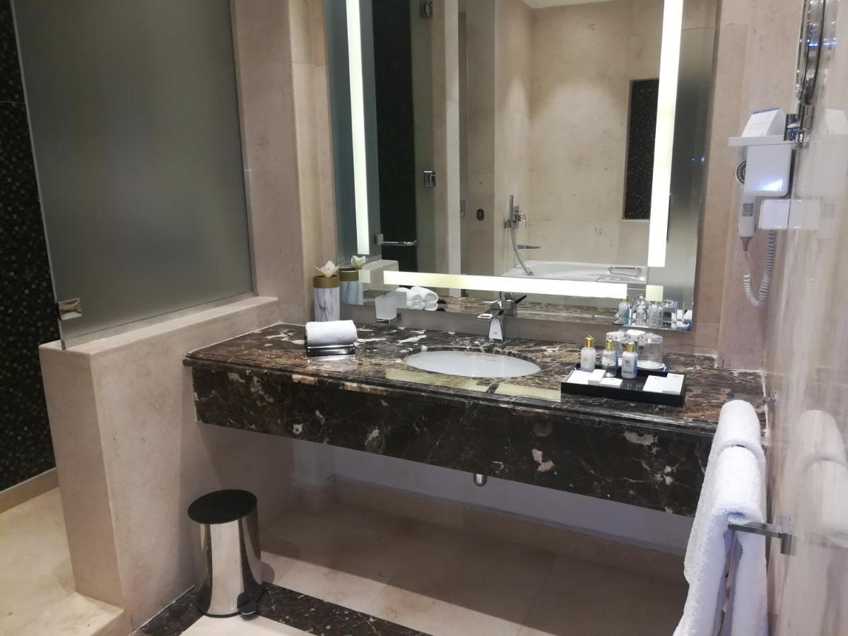 톨립 리조트 엘 갈라라 마제스틱 아인 수크나 외부 사진 The photo shows a modern bathroom with elegant design features. There is a large vanity made of dark marble with a sink and a set of toiletries neatly arranged on top. A mirror with built-in lighting is mounted above the sink. To the side, there is a