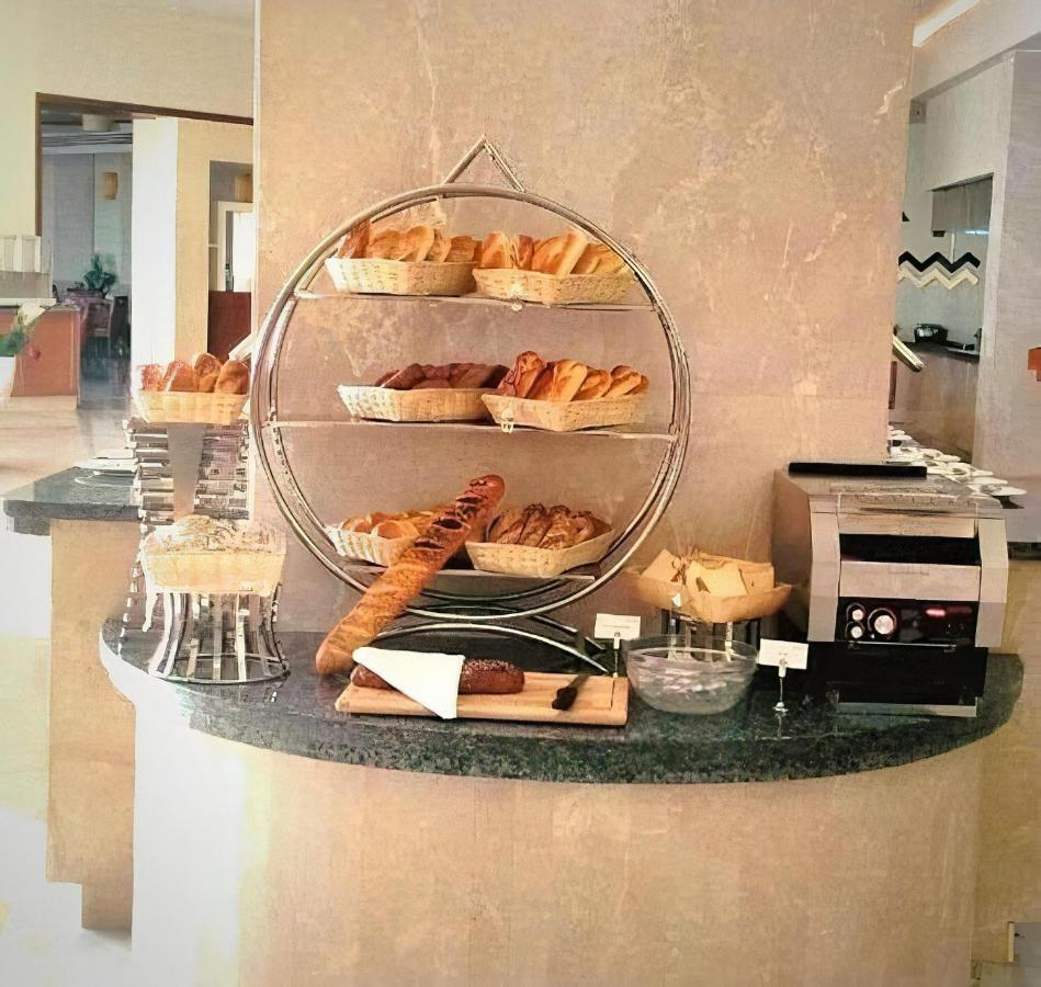 톨립 리조트 엘 갈라라 마제스틱 아인 수크나 외부 사진 The photo shows a display of various types of bread and pastries arranged on a countertop. There are several baskets stacked on a circular stand, filled with different baked goods like croissants and rolls. A long baguette is placed horizontally on a
