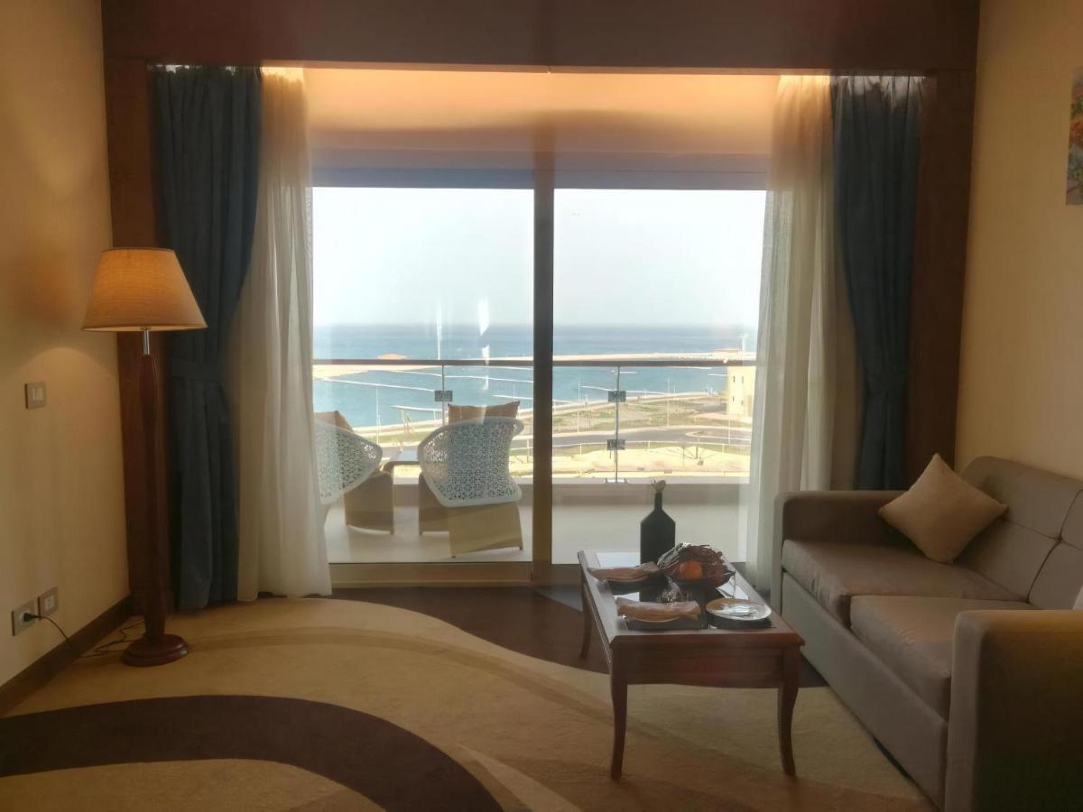 톨립 리조트 엘 갈라라 마제스틱 아인 수크나 외부 사진 The photo shows a stylish interior of a room with a large window overlooking the sea. The window features sheer and thick curtains and is framed by a balcony with two chairs visible outside. Inside, there is a comfortable-looking sofa, a coffee table