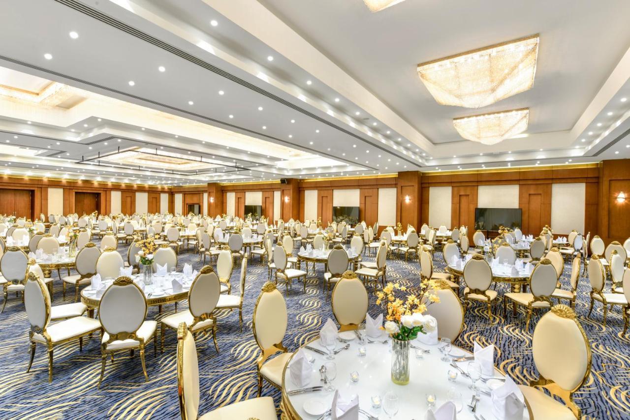 톨립 리조트 엘 갈라라 마제스틱 아인 수크나 외부 사진 The photo shows a spacious and elegantly decorated banquet hall. The room features numerous round dining tables, each set with white tablecloths and adorned with decorative centerpieces, likely flowers. The chairs surrounding the tables have a luxuri